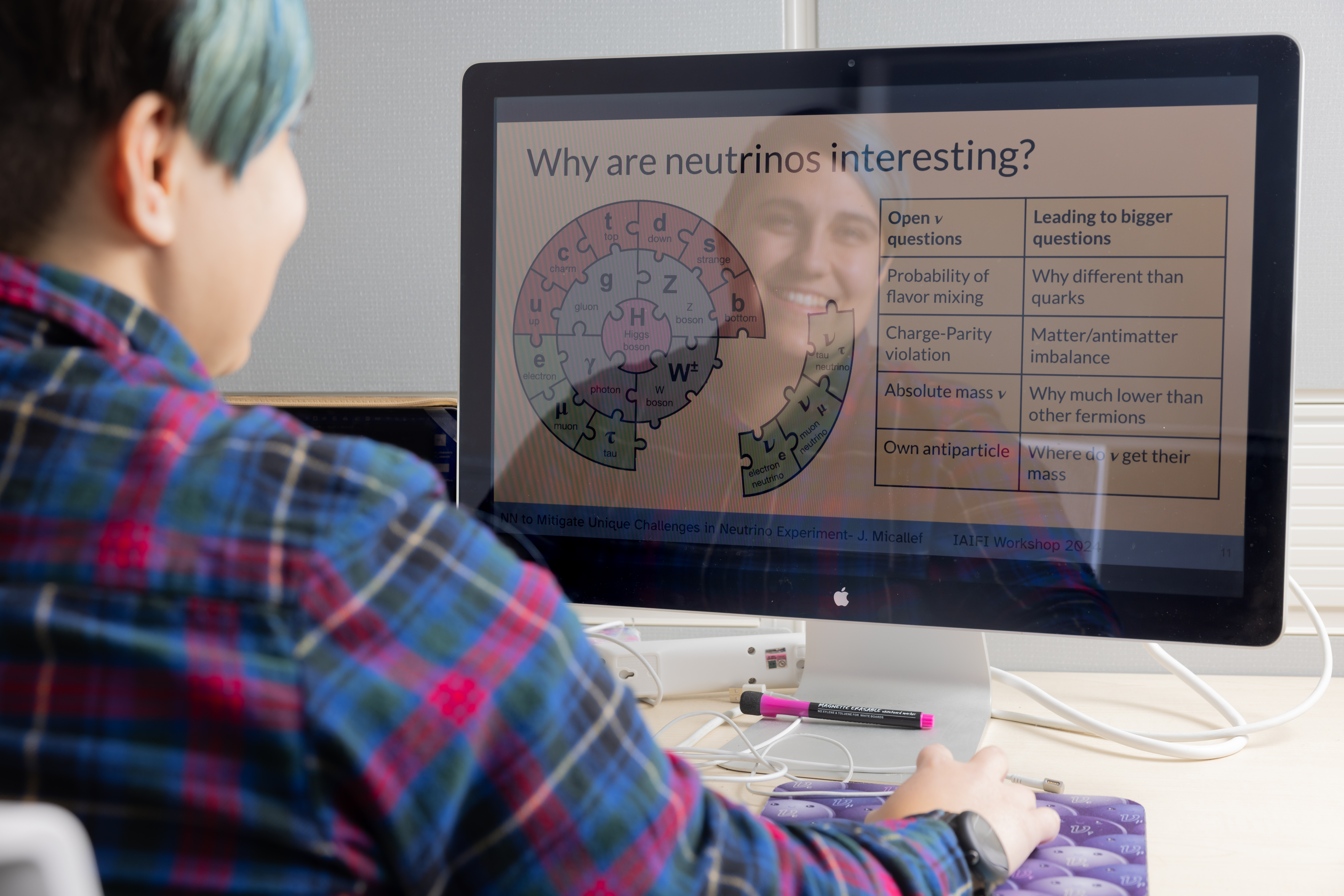 Jessie sitting at their computer with their reflection visible over the graphic they created to explain the standard model of particle physics